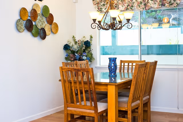 A dining room with a wooden table and six matching wooden chairs with cushioned seats. A blue decorative vase is placed on the table. On the wall to the left, there is a decorative arrangement of colorful plates. A floral curtain hangs above a large window letting in natural light. A chandelier with six lights hangs above the table. A floral arrangement is placed in the corner near the window.