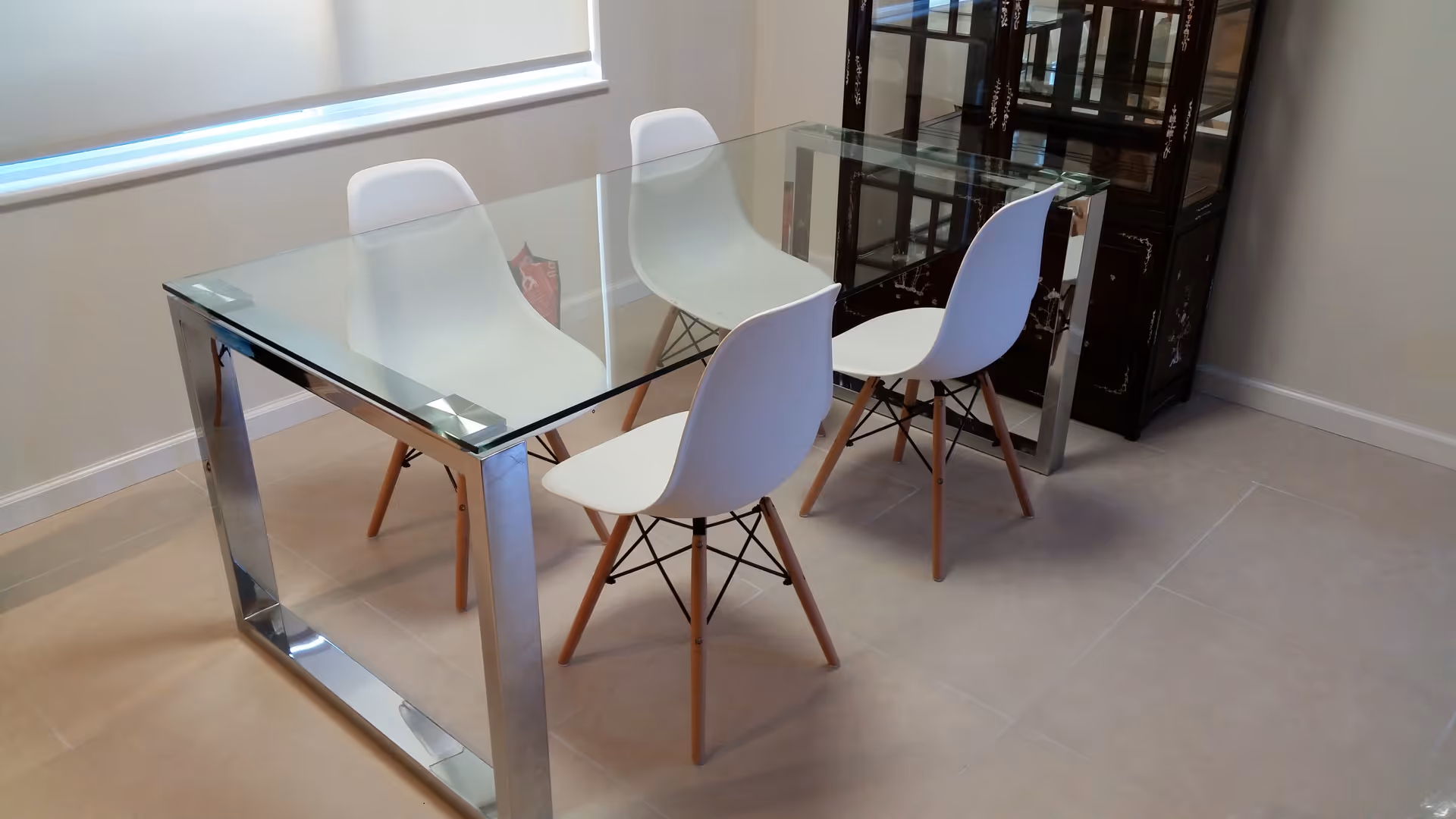 A modern dining area with a glass-top table and four white chairs with wooden legs, positioned near a window with a white blind. There is a dark wooden cabinet with glass doors in the corner of the room.