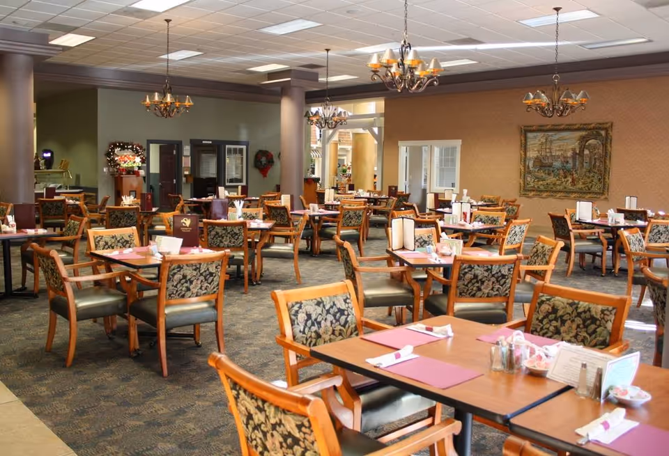 A spacious dining room with multiple wooden tables and chairs arranged neatly. Each table has placemats, napkins, and condiments. The room features chandeliers hanging from the ceiling, a carpeted floor, and a tapestry on the wall. The atmosphere is warm and inviting.