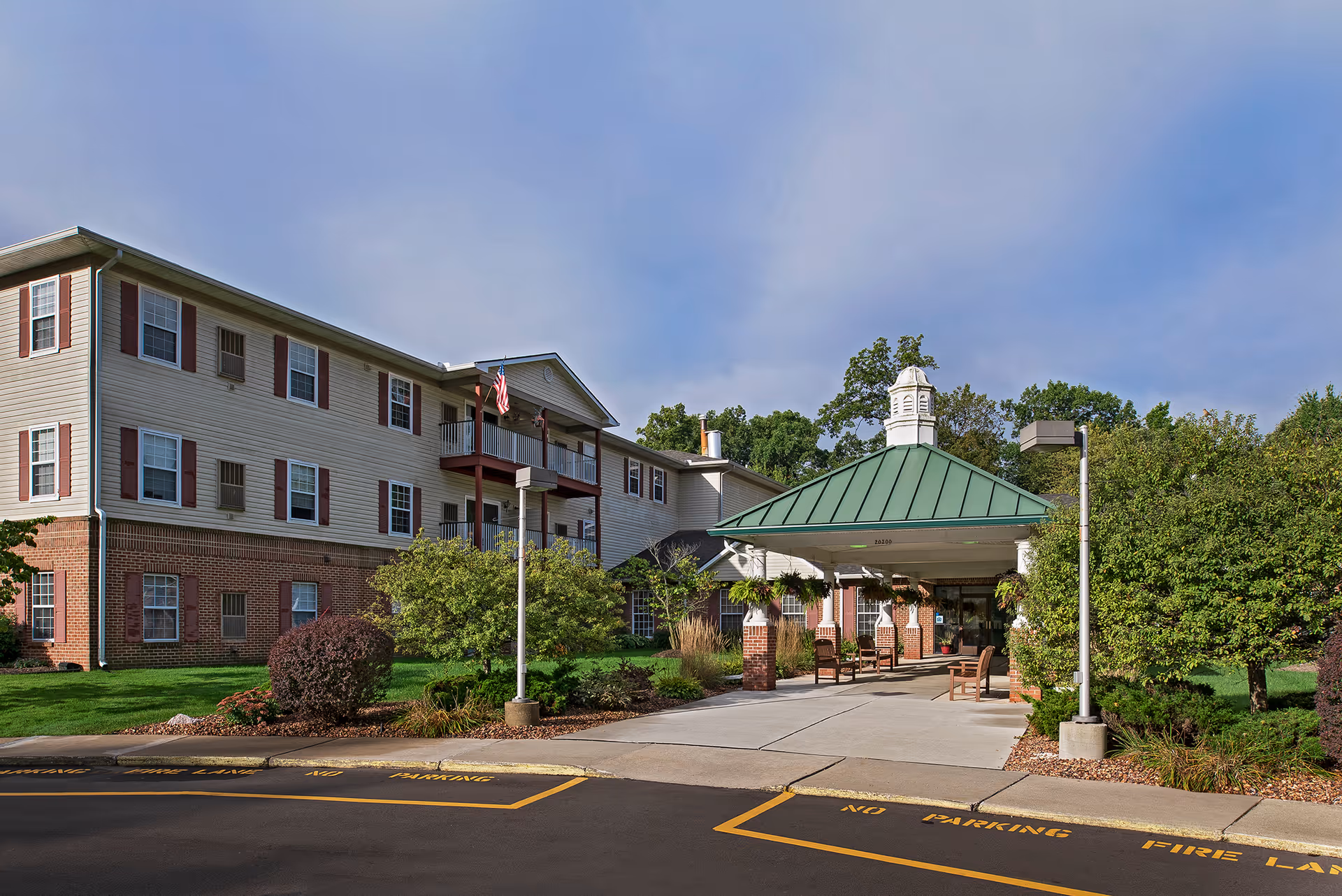 Exterior view of a senior living facility building with a covered entrance featuring a green metal roof and brick pillars. The building has three stories with beige siding and red shutters on the windows. There are trees, shrubs, and a well-maintained lawn surrounding the entrance area. The sky is clear with some clouds.