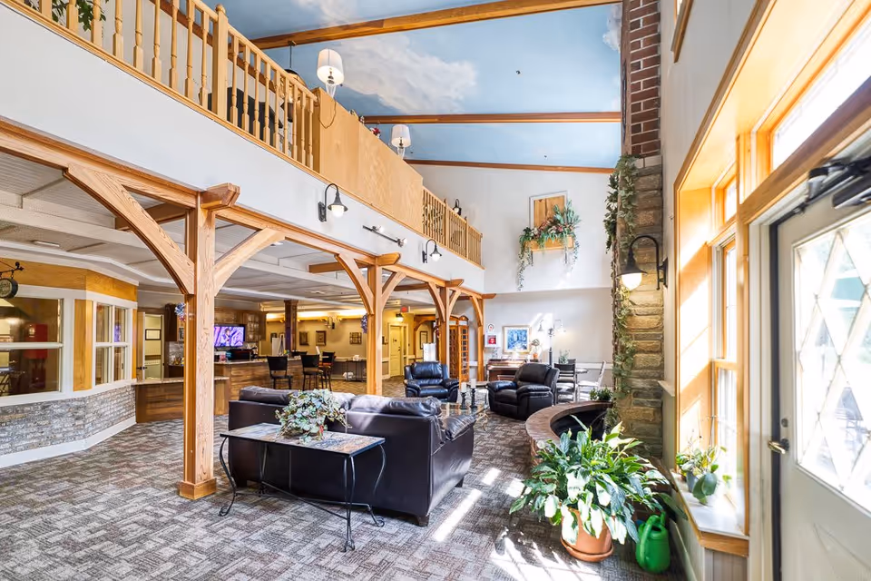 A bright and spacious living room area in Saint Therese of Westlake featuring leather sofas and armchairs arranged around a coffee table. The room has large windows letting in natural light, wooden beams and railings, indoor plants, and a high ceiling painted with a sky mural. There is a reception or dining area visible in the background.