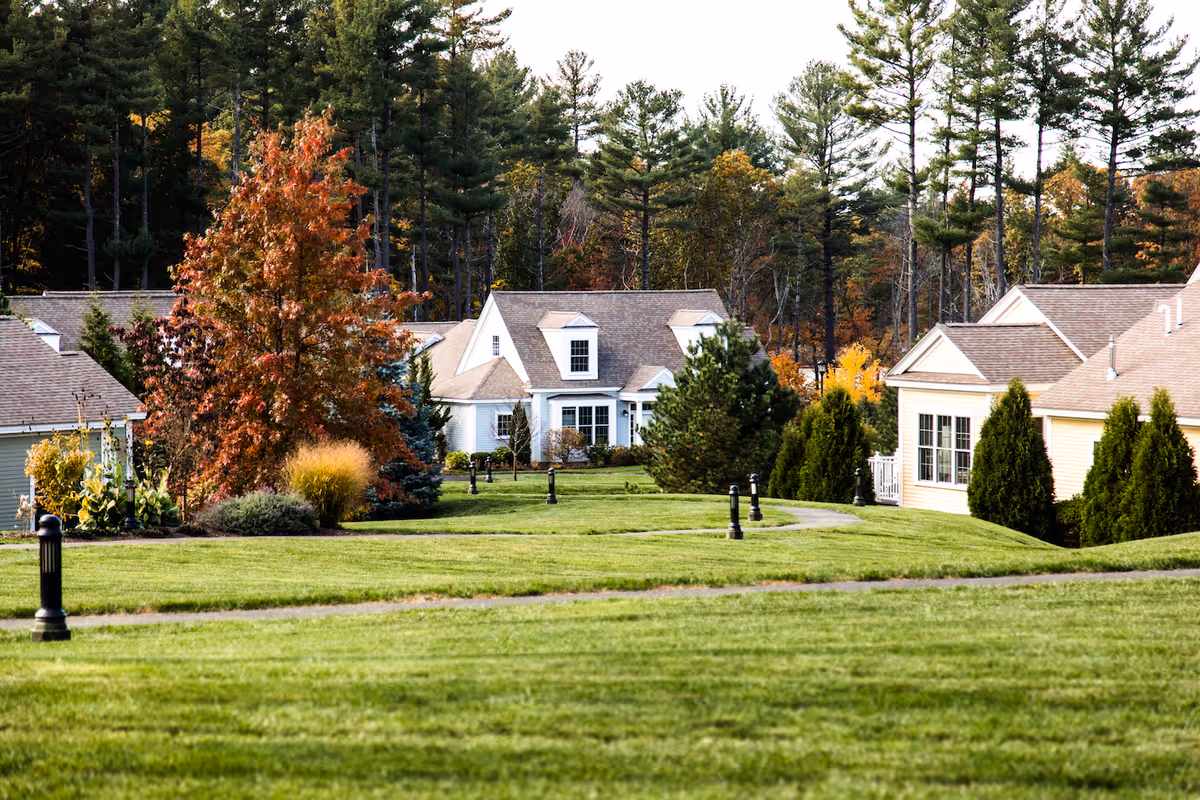 A scenic view of a senior living community with well-maintained green lawns, paved walkways, and several single-story houses surrounded by trees with autumn foliage.