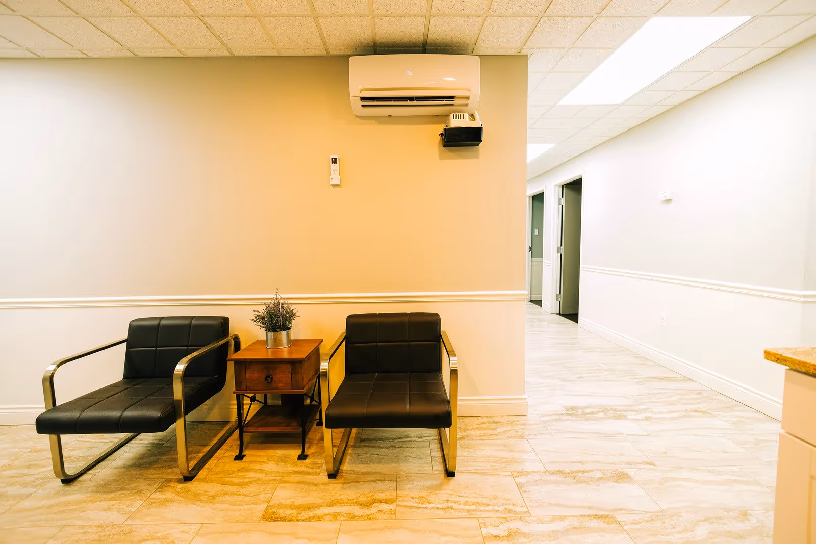 A hallway area in The Vernon Center with two black leather chairs with metal armrests separated by a small wooden side table holding a potted plant. The walls are painted beige and white with a white chair rail, and there is a white air conditioning unit mounted on the wall above the chairs. The floor is covered with light-colored tiles, and the hallway extends to the right with several doorways visible.