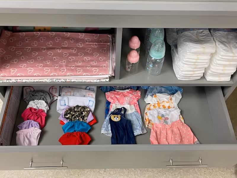 An open drawer containing neatly folded baby clothes, diapers, baby bottles, and pink patterned blankets on a shelf above the drawer.