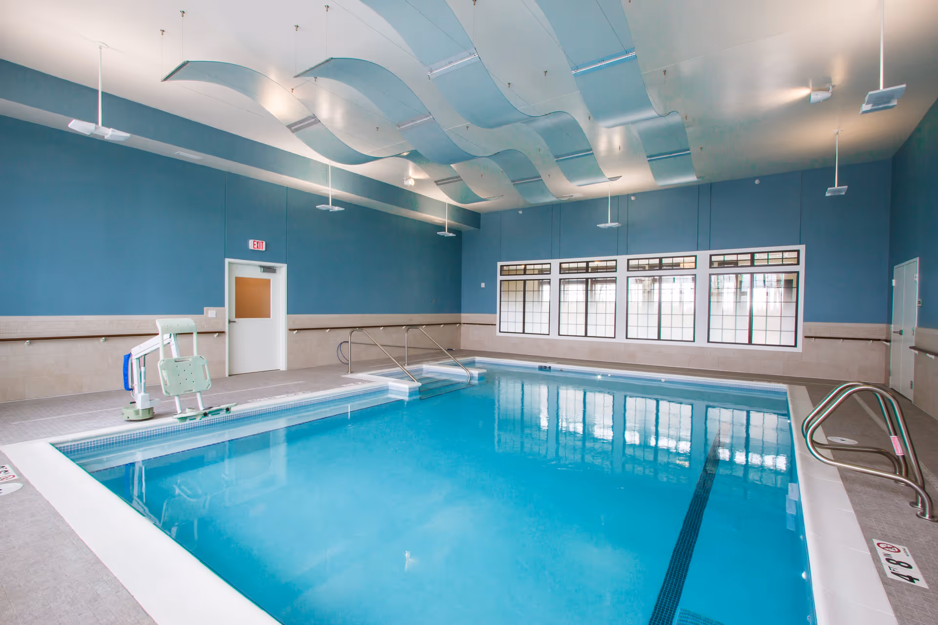 Indoor swimming pool with clear blue water, light blue walls, and large windows. The ceiling features decorative wave-like panels and several hanging lights. There is a pool lift chair on the left side and metal handrails on the right side of the pool.