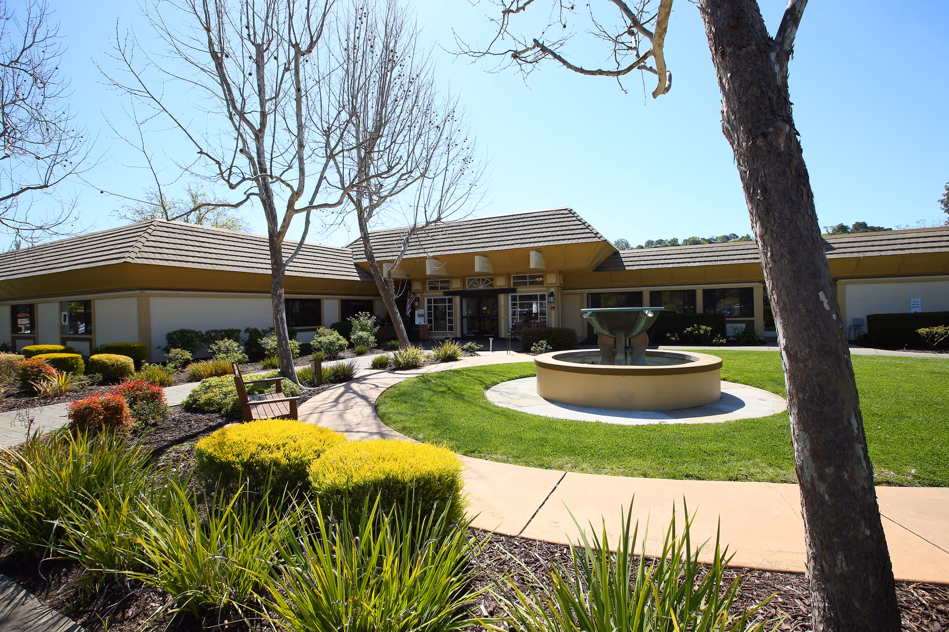 Exterior view of Walnut Creek Skilled Nursing & Rehabilitation Center featuring a circular fountain in the center of a well-maintained lawn, surrounded by a curved walkway, bushes, and trees with a clear blue sky above.