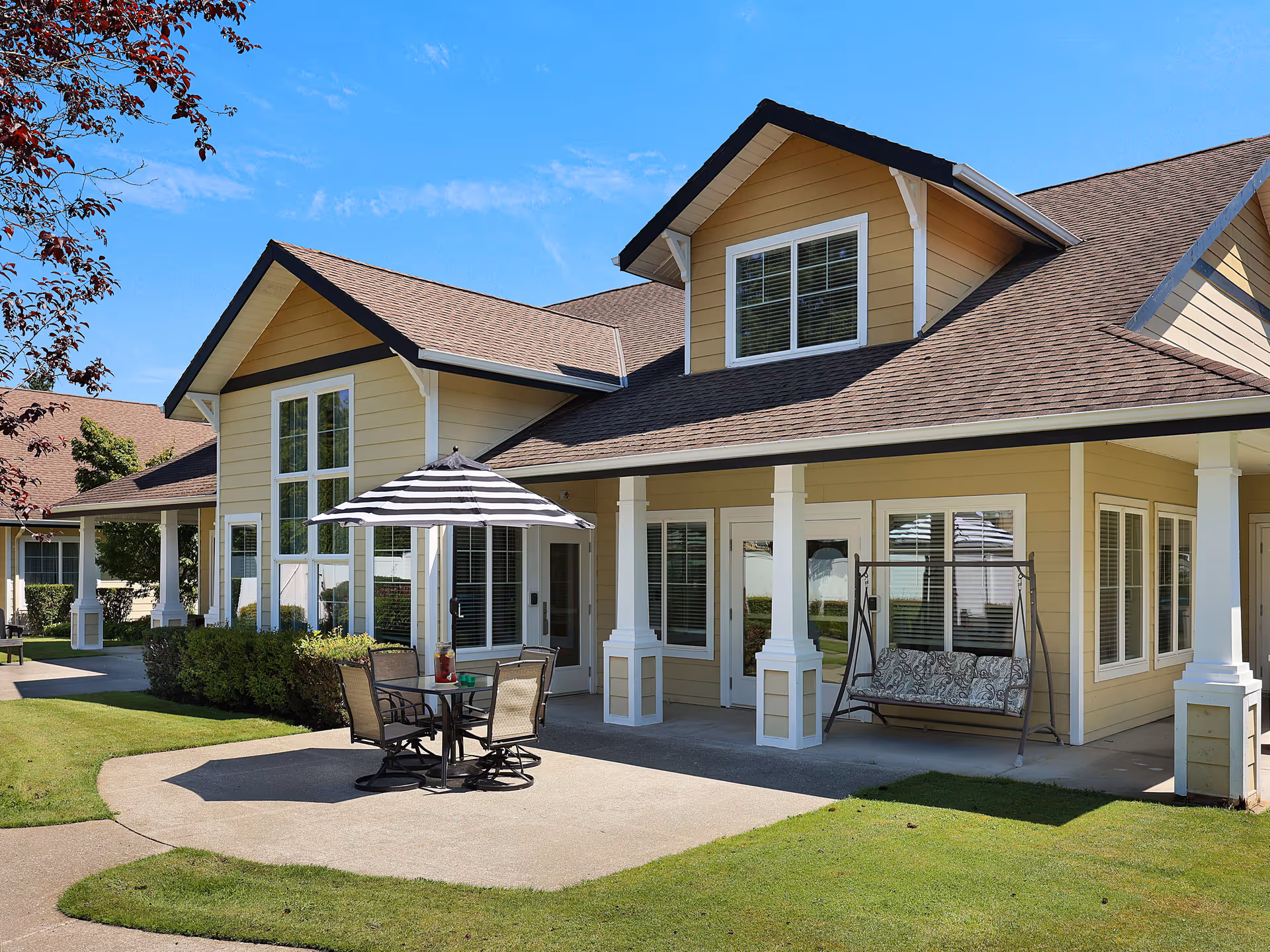 Exterior view of a yellow two-story building with white trim and a brown roof under a clear blue sky. The building has large windows and a covered patio area with white columns. On the patio, there is a table with four chairs and a striped umbrella, as well as a cushioned swing. The surrounding area has green grass and some bushes.