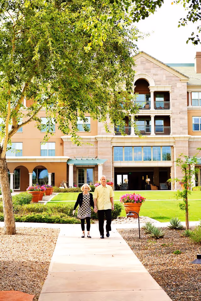 An elderly couple walking hand in hand on a paved pathway in front of a multi-story senior living facility building with large windows and balconies. The area is landscaped with green grass, trees, and flower pots with colorful flowers.