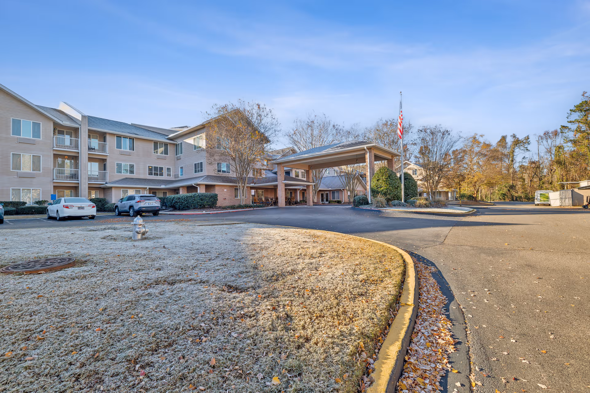 Exterior view of a multi-story senior living facility building with a covered entrance, several parked cars, a fire hydrant, and a flagpole with an American flag. The surrounding area includes frosty grass, trees, and a clear blue sky.