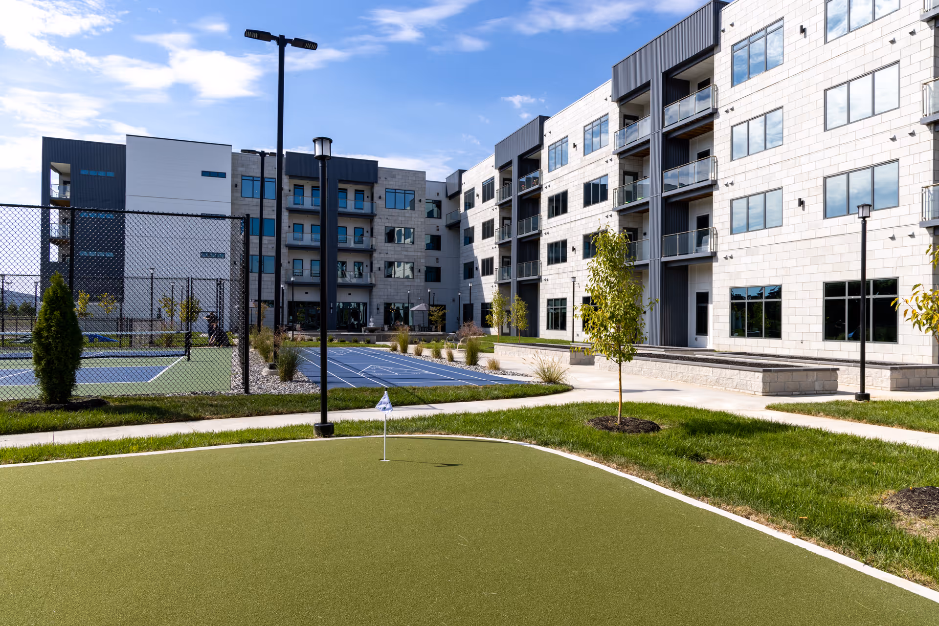 Outdoor view of a modern senior living facility with a small putting green in the foreground, a tennis court enclosed by a fence, and a multi-story building with balconies and large windows under a partly cloudy sky.