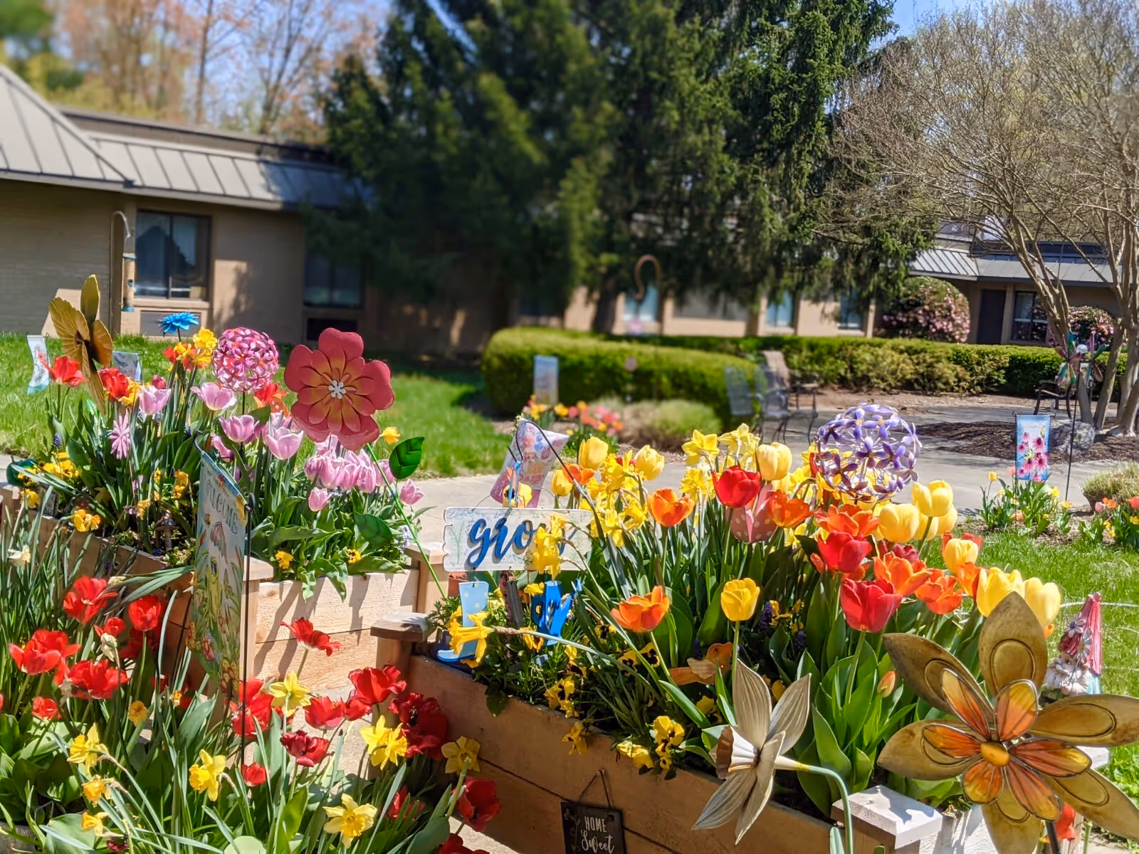 A colorful garden with various blooming flowers including tulips and daffodils planted in wooden raised beds. Decorative garden ornaments such as metal flowers and butterflies are placed among the plants. In the background, there is a building with windows and a metal roof, surrounded by green bushes and trees under a clear sky.