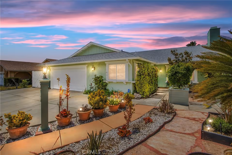 Front exterior of a single-story house with a garage, driveway, potted plants, and a landscaped walkway at dusk.