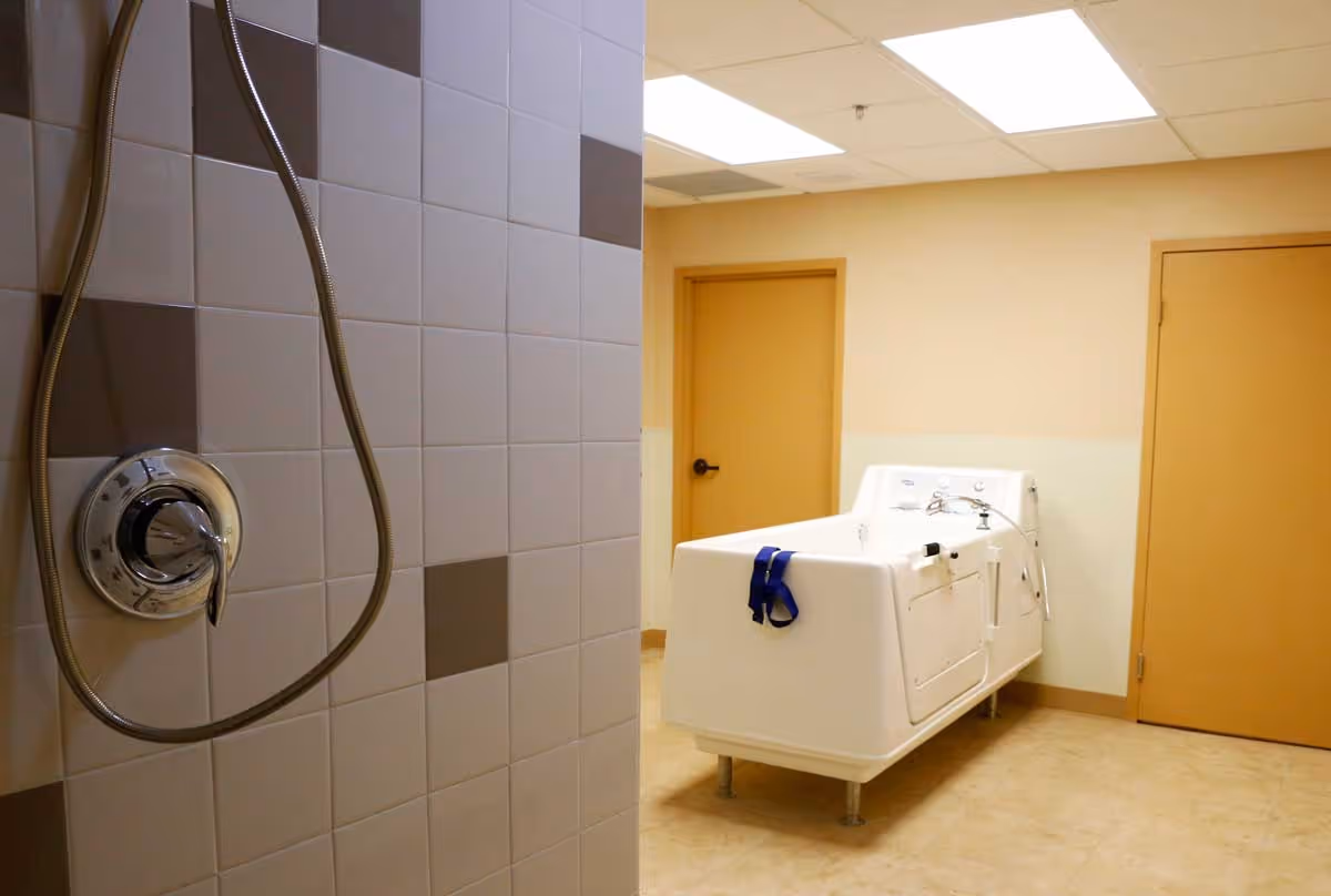 A tiled shower wall with a metal shower handle and hose in the foreground, and a white accessible bathtub with blue straps in a beige room with two closed doors in the background.