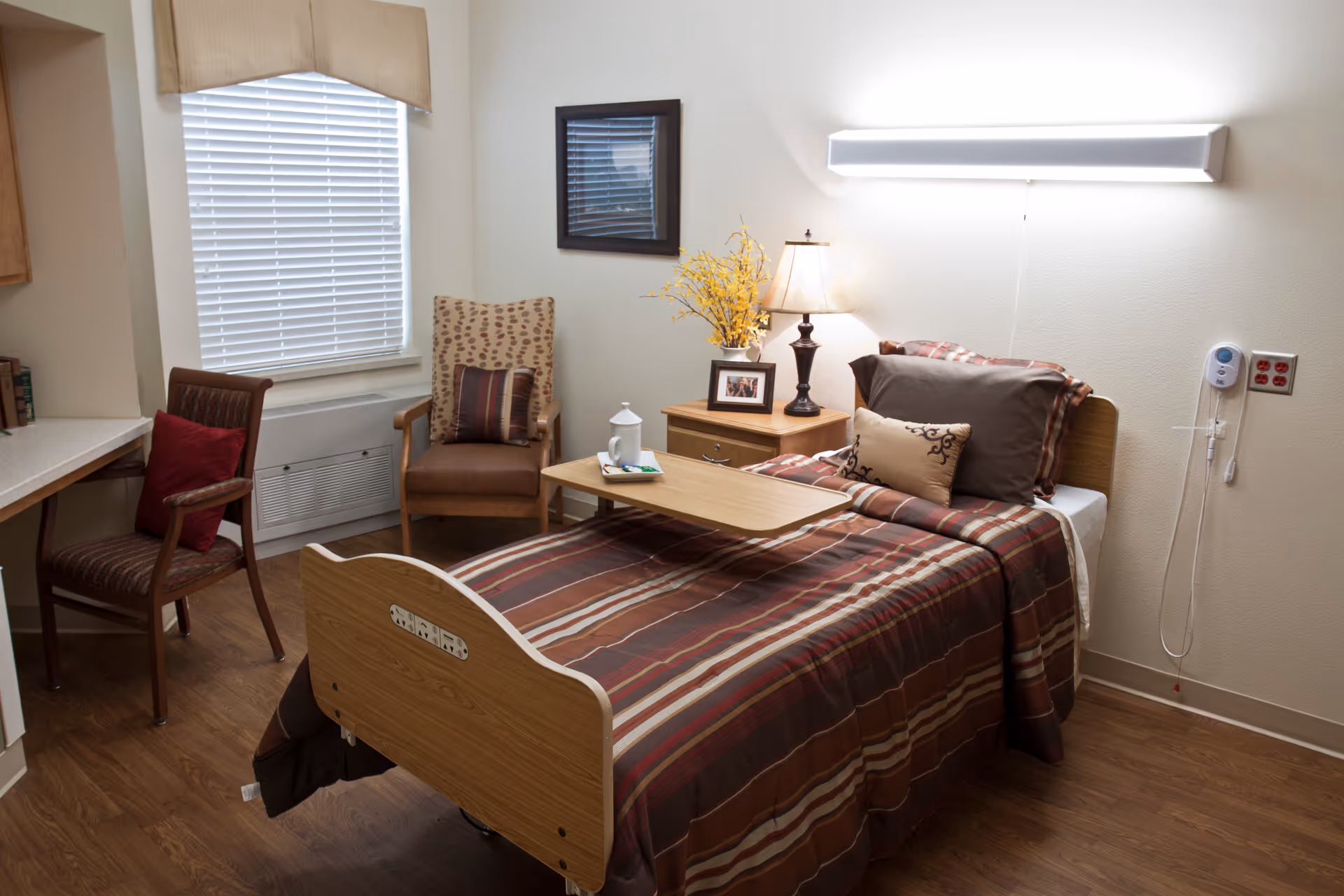 A neatly furnished senior living bedroom with a single adjustable bed, bedside table with a lamp, and two chairs by a window.