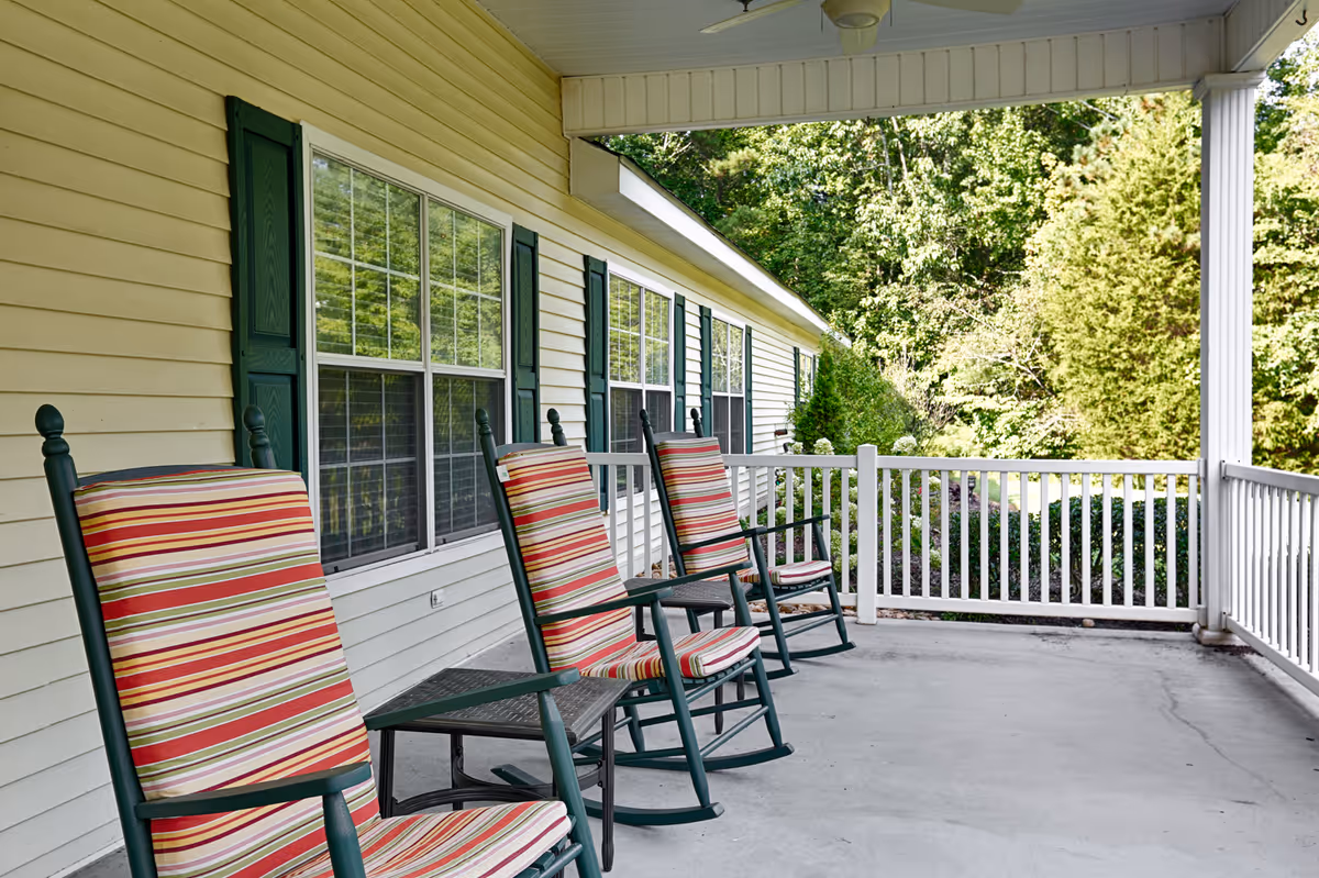 A covered porch with four green rocking chairs featuring red, white, and green striped cushions. The porch has white railings and overlooks a green garden area with trees and bushes. The building exterior is light yellow with green window shutters.
