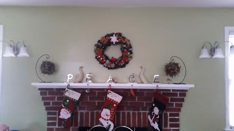 A brick fireplace decorated for Christmas with three stockings hanging from the mantel. On the mantel, there are decorative letters spelling 'PEACE', two small bird figurines, two hanging wreaths on either side, and a larger Christmas wreath centered above. The wall behind is painted light green, and there are two wall-mounted light fixtures with white shades on either side of the fireplace.