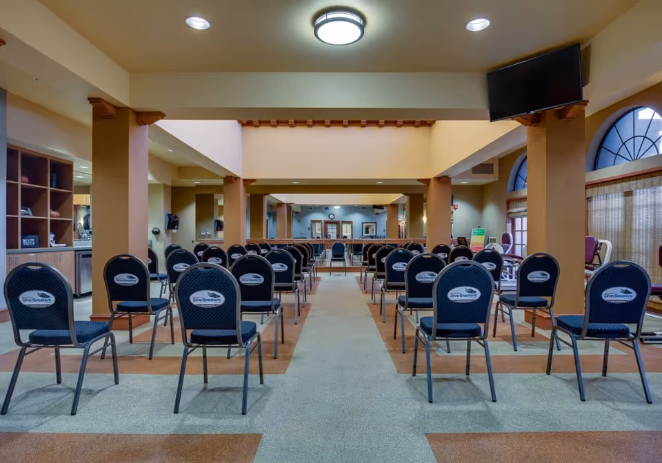 A spacious room set up with rows of chairs facing a mirrored wall. The chairs have labels that say 'SilverSneakers'. The room has beige walls, a high ceiling with recessed lighting, and large arched windows with curtains. There is a TV mounted on the right wall and exercise equipment visible in the background.