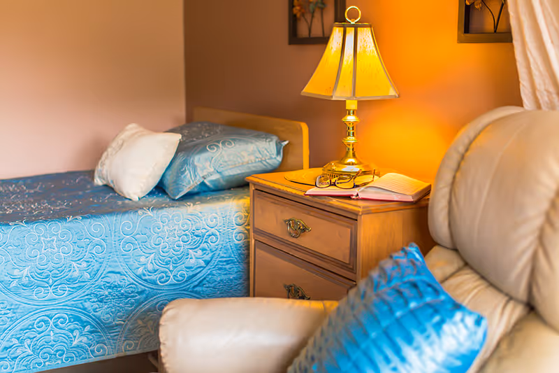 A cozy bedroom featuring a single bed with a blue patterned bedspread and white pillow, a wooden nightstand with a brass lamp, an open book, and a pair of glasses. In the foreground, there is a beige leather armchair with a blue cushion.