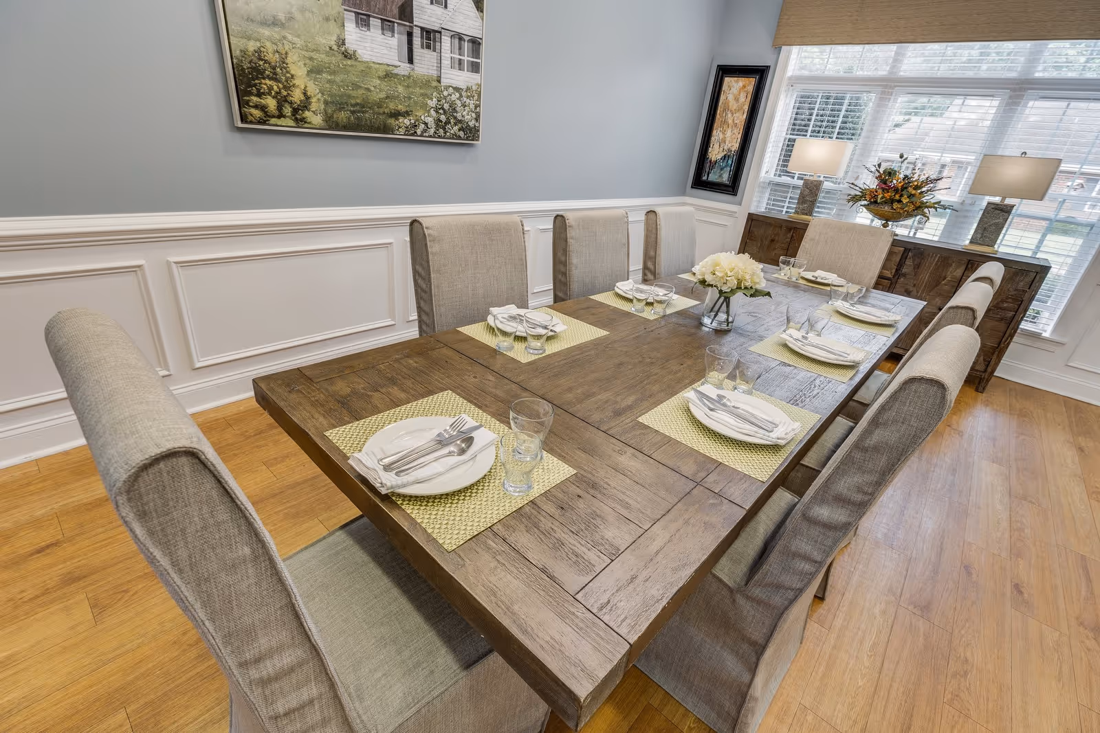 A dining room with a large wooden table set for six people. Each place setting includes a white plate, silverware wrapped in a napkin, and a glass. The chairs are upholstered in a light beige fabric. The room has light blue walls with white wainscoting, a wooden floor, and a sideboard with two lamps and a floral arrangement in front of a large window with white blinds. Two framed paintings hang on the walls.