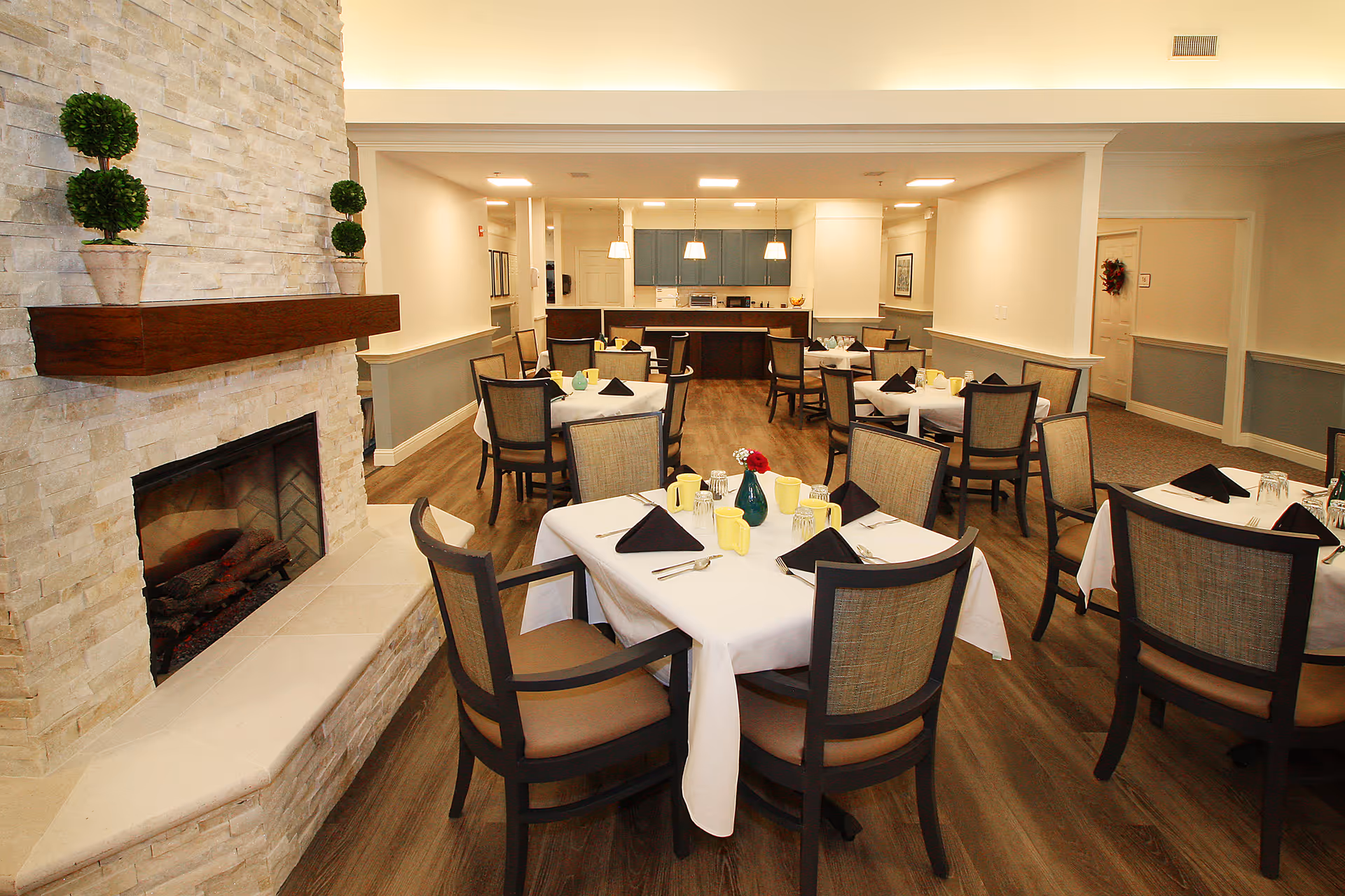 Dining room with multiple tables set with white tablecloths, chairs, and a stone fireplace in a senior living facility.
