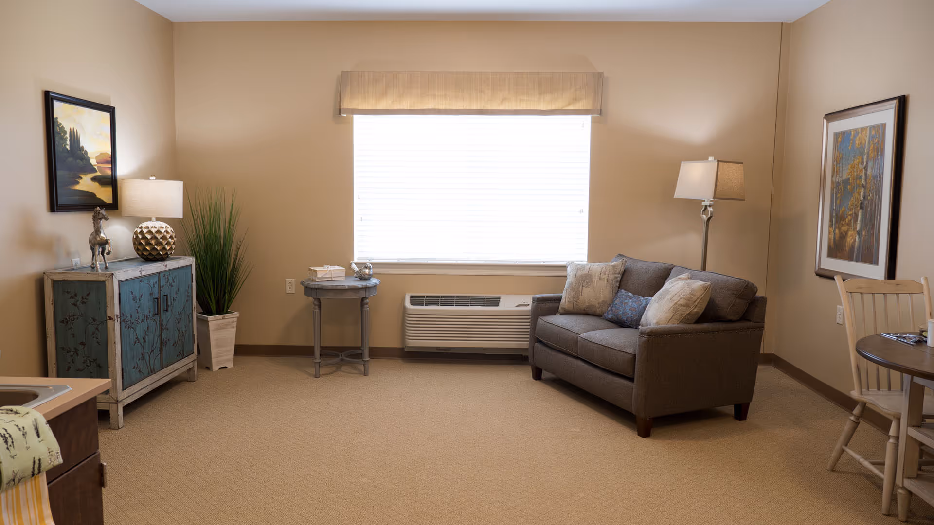 Well-lit living room with a loveseat, side tables, a decorative cabinet, and a small dining area in front of a large window.