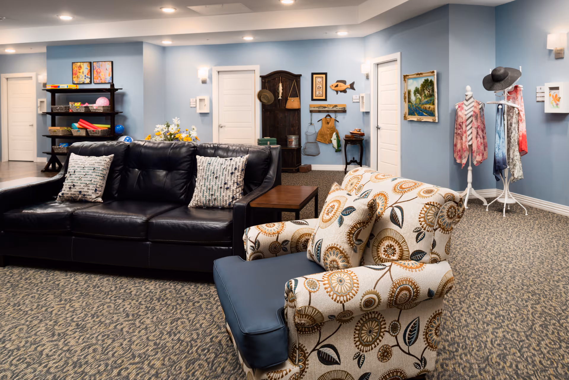 A cozy senior living common area with a black leather sofa and a patterned armchair with cushions. The room has light blue walls, carpeted floor, and several doors. There is a wooden shelf with baskets and colorful items, a coat rack with hats and scarves, framed artwork on the walls, and a small table with flowers.