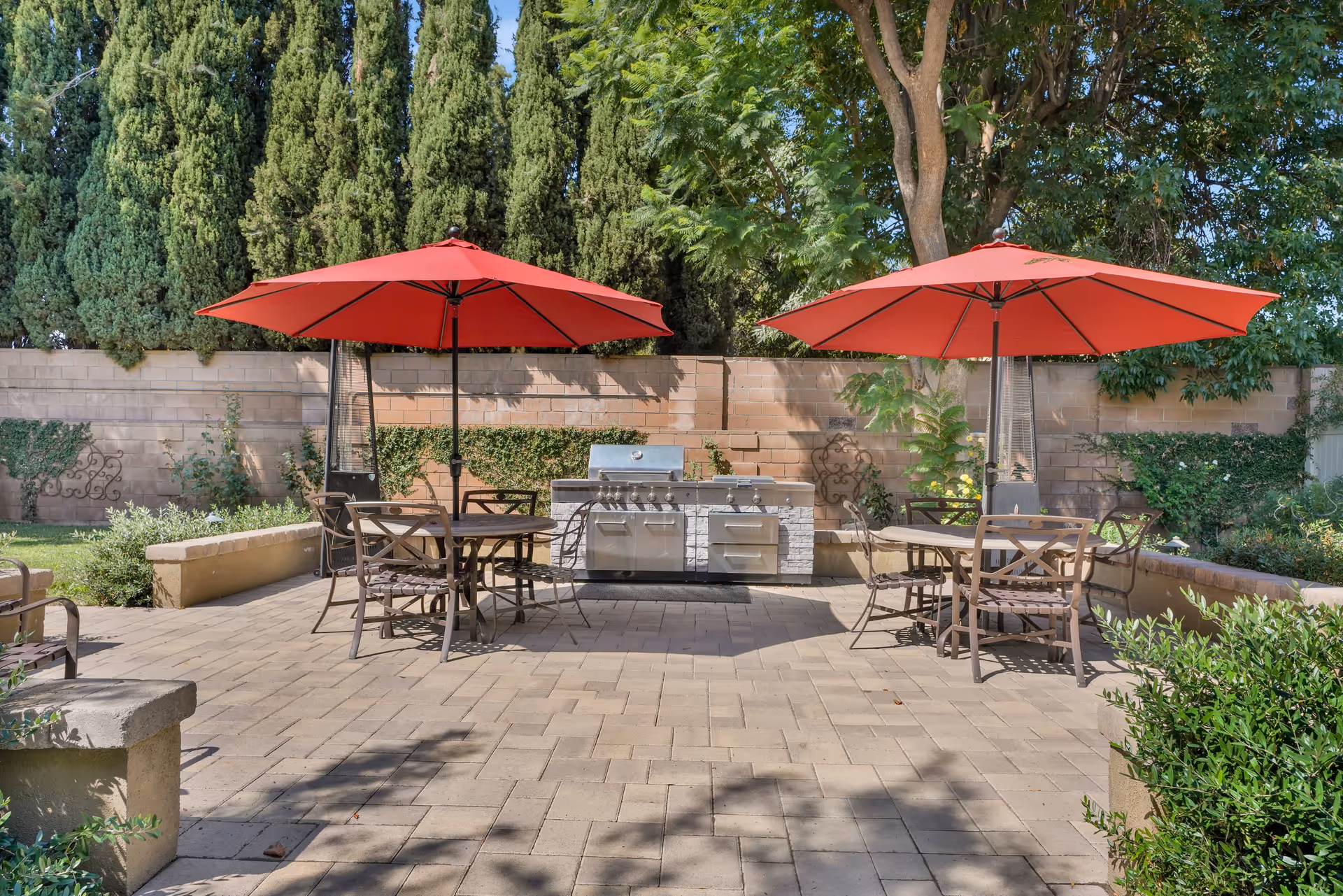 Outdoor patio area with two round tables, each shaded by a large red umbrella. Each table is surrounded by metal chairs. In the background, there is a built-in stainless steel grill against a brick wall, with tall green trees and plants surrounding the area.