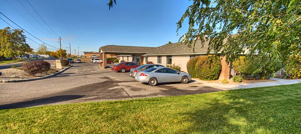 Exterior view of a single-story residential care facility with a parking lot in front containing several parked cars. The building is surrounded by green grass, bushes, and trees under a clear blue sky.