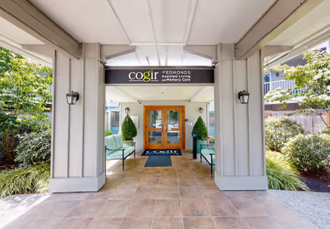 Covered entrance area of Cogir of Edmonds Assisted Living and Memory Care facility with double wooden doors, two green cushioned benches, potted plants, and a sign above the entrance displaying the facility name. Surrounding greenery and outdoor landscaping are visible.