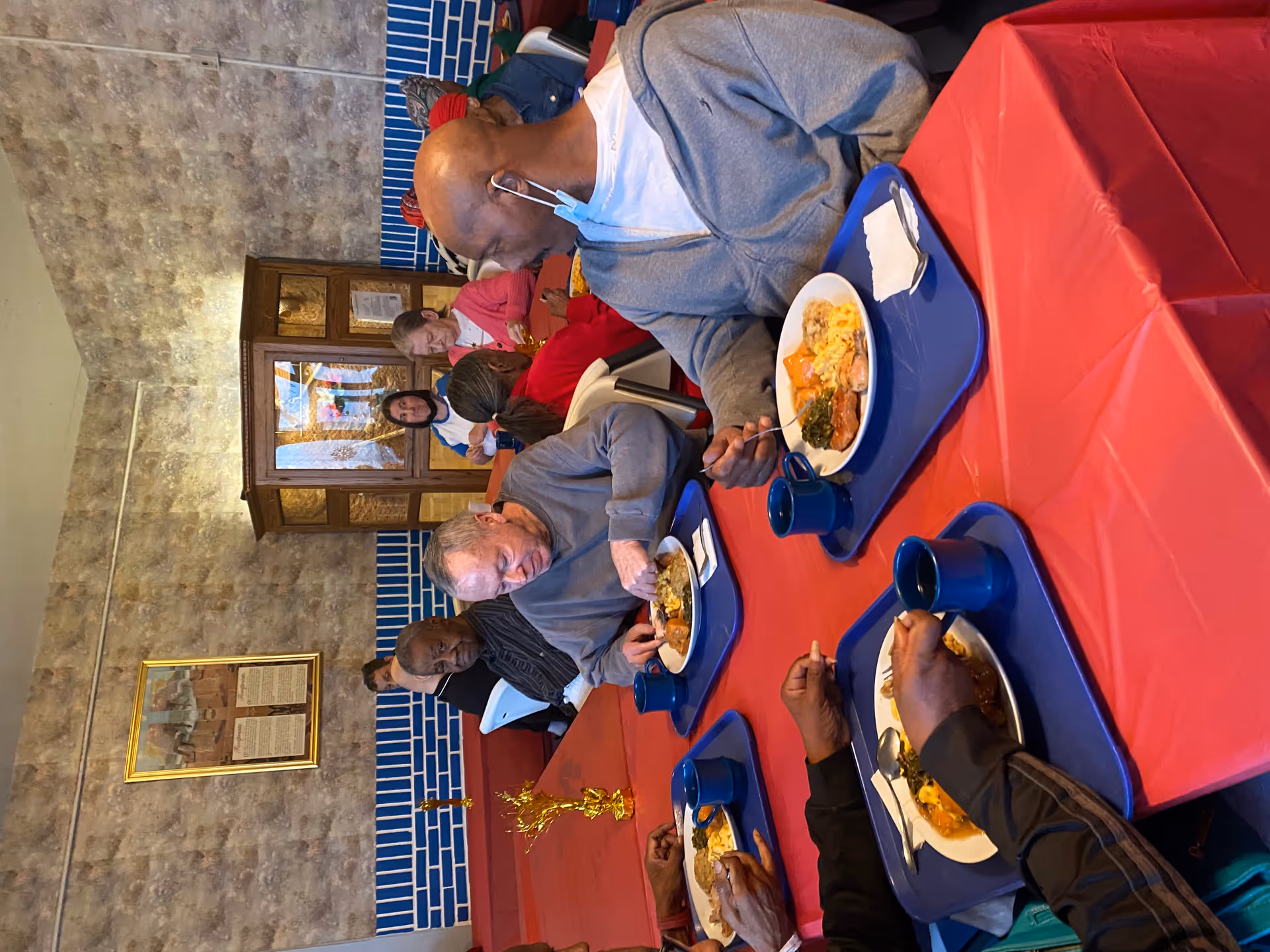 Several elderly residents sit at a red-covered table in a communal dining room eating meals on blue trays.