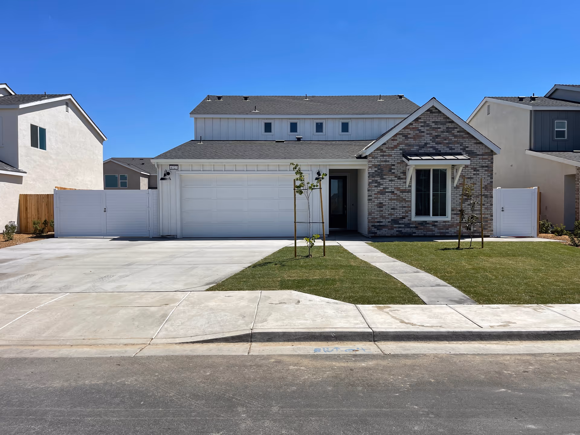 Front exterior of a modern suburban house with a two-car garage, driveway, lawn, and young trees under a clear blue sky.