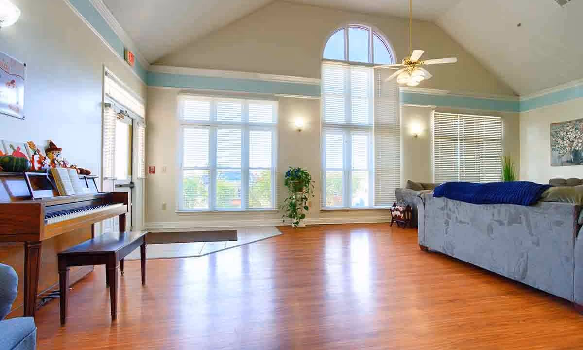 A spacious living room with large windows allowing natural light to fill the room. The room features a wooden floor, a piano with a bench on the left side, a ceiling fan with lights, a gray couch with a blue throw blanket, a plant near the windows, and a painting on the wall.