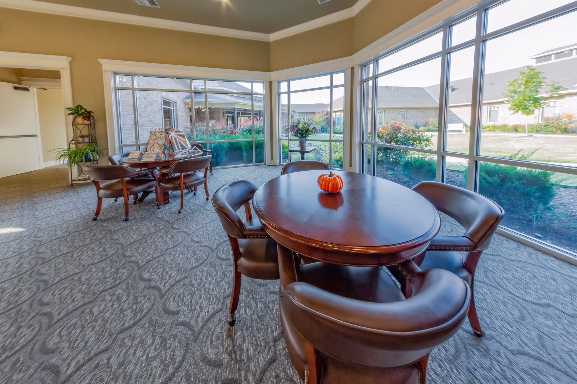 A bright room with large windows overlooking a garden area. Inside, there are two round wooden tables surrounded by brown leather chairs. One table has a small pumpkin on it, and the other has decorative items and plants nearby. The room has patterned carpet and beige walls with white trim.
