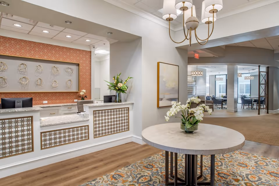 Reception area of Brookdale Galleria featuring a white and gold front desk with two computer monitors, decorative wall art behind the desk, a round table with a flower arrangement in the foreground, and a seating area with tables and chairs visible in the background.