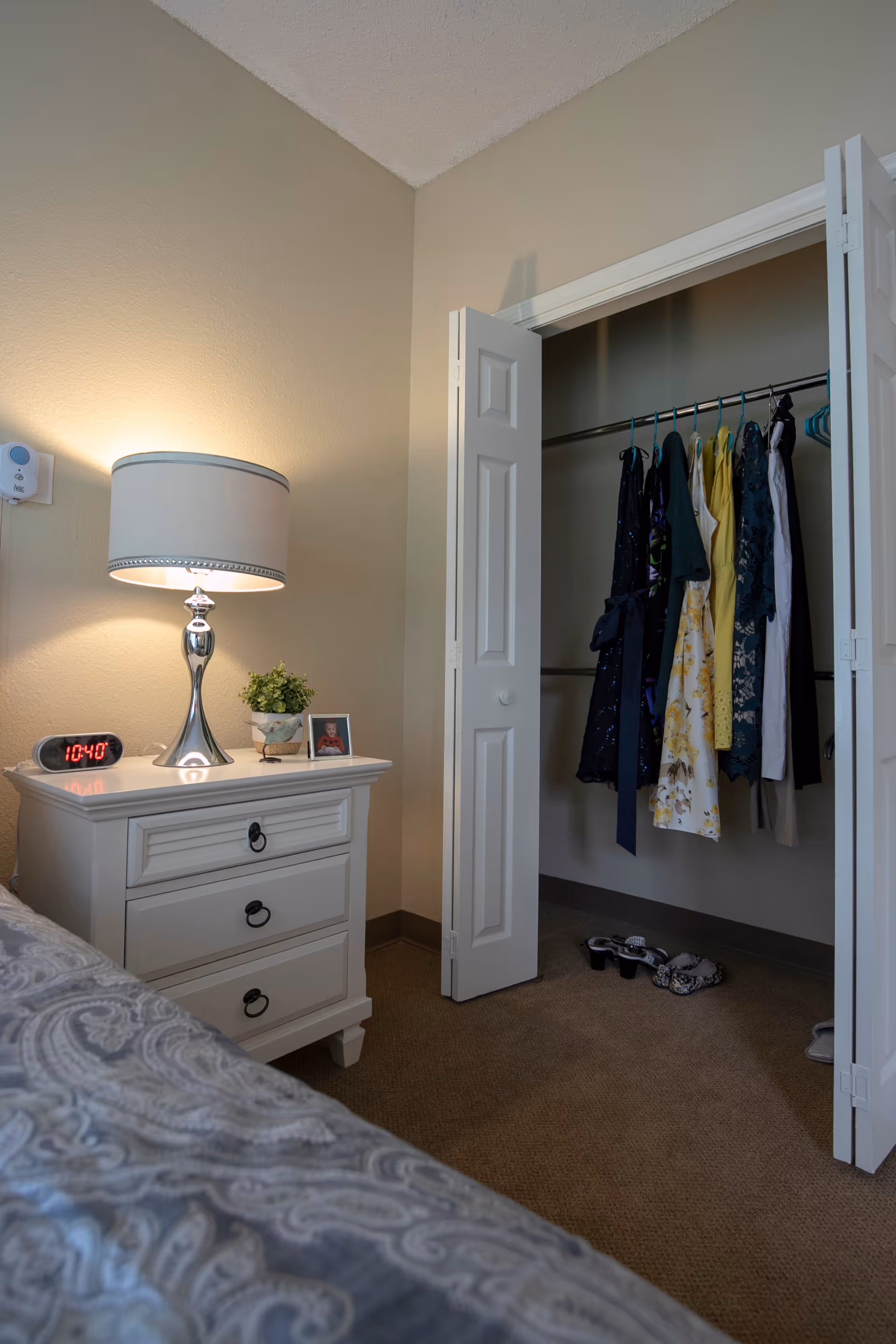 A bedroom corner featuring a white nightstand with three drawers, a silver lamp with a white shade, a digital alarm clock showing 10:40, a small potted plant, and a framed photo. Next to the nightstand is an open closet with white bi-fold doors, containing several hanging dresses and a pair of shoes on the carpeted floor.