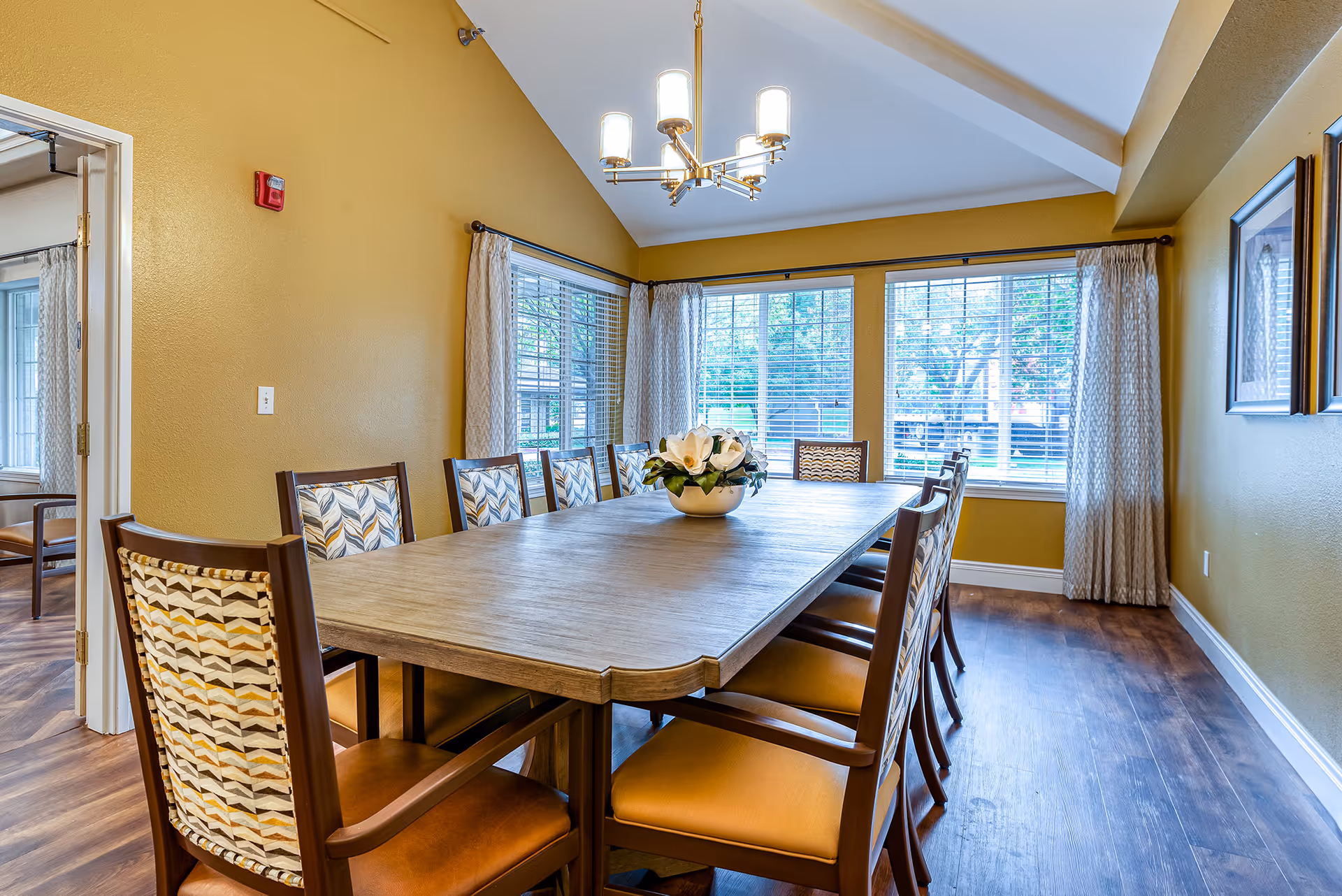 A bright dining room with a long wooden table surrounded by ten chairs with patterned backs and cushioned seats. A white floral centerpiece sits on the table. The room has mustard yellow walls, large windows with white blinds and light-colored curtains, and a modern chandelier hanging from a vaulted ceiling. Hardwood flooring completes the space.