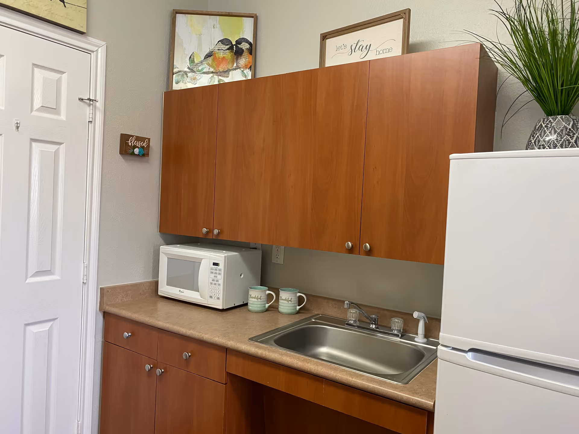 A small kitchen area with wooden cabinets, a white microwave on the countertop, two mugs, a stainless steel sink, and a white refrigerator. There are framed pictures and a small plant on top of the cabinets.