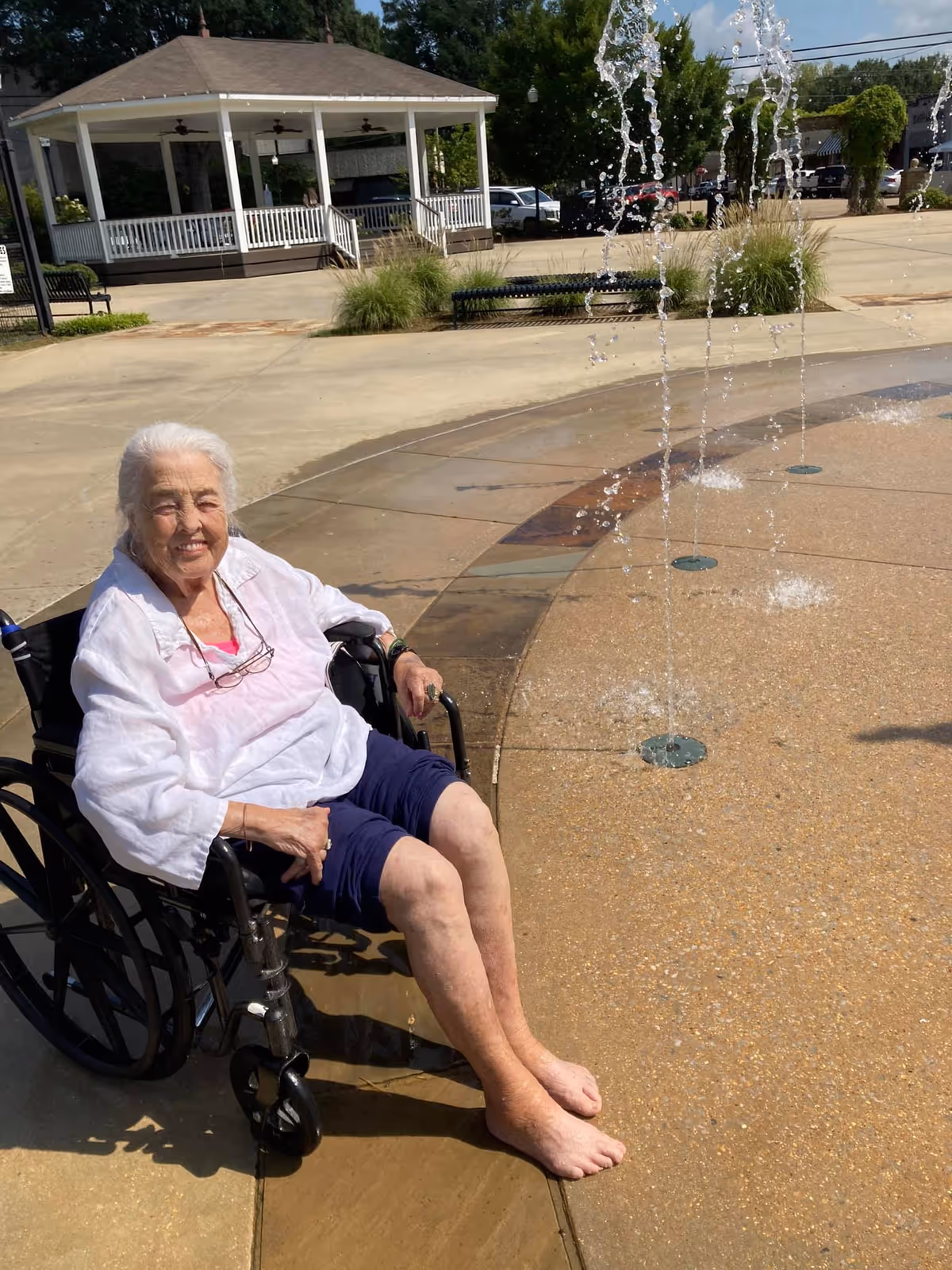 An elderly woman sits in a wheelchair with her feet in a water fountain at a senior living facility.