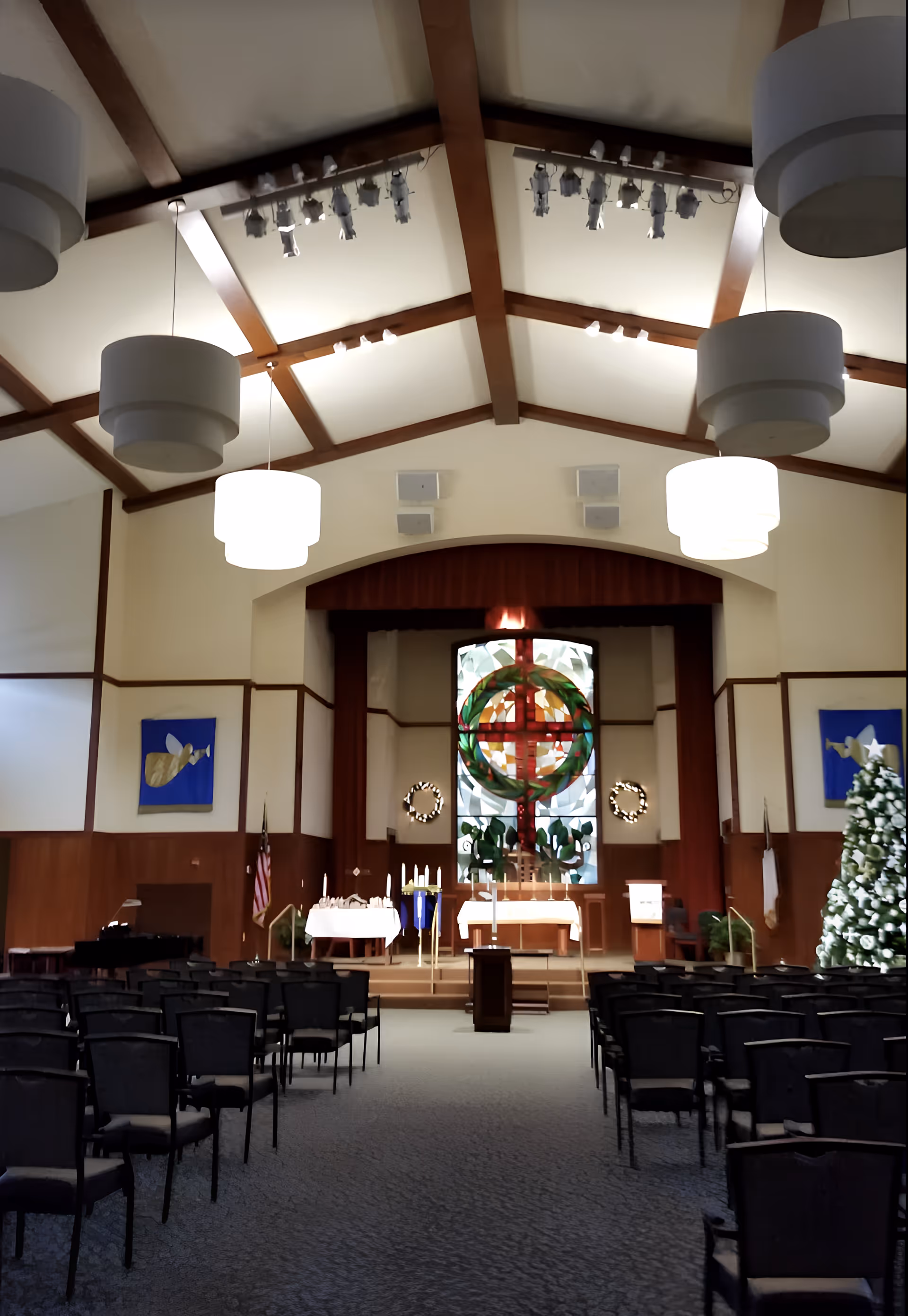 Interior view of a chapel with rows of chairs facing an altar. The altar area features a large stained glass window with a circular wreath design and a red cross. There are candles on stands and tables with white cloths. The ceiling has wooden beams and hanging round light fixtures. A decorated Christmas tree is visible on the right side of the chapel.