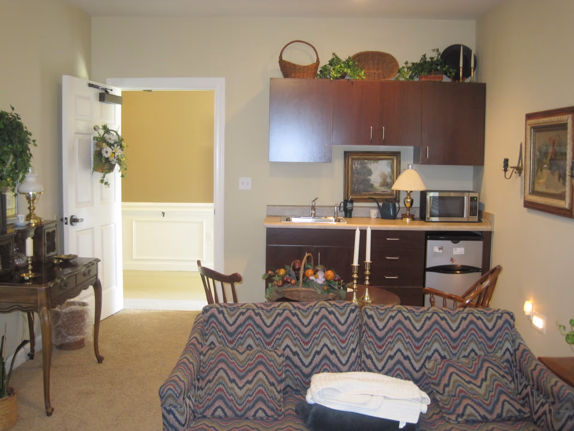 Interior view of a cozy assisted living facility room featuring a small kitchenette with dark wood cabinets, a sink, microwave, and mini fridge. In front of the kitchenette is a patterned sofa with folded blankets on it, a small round table with decorative fruit and two candlesticks, and wooden chairs. The room has beige walls with framed artwork, a side table with a lamp and decorative items, and an open door leading to another room with light-colored walls and carpet.