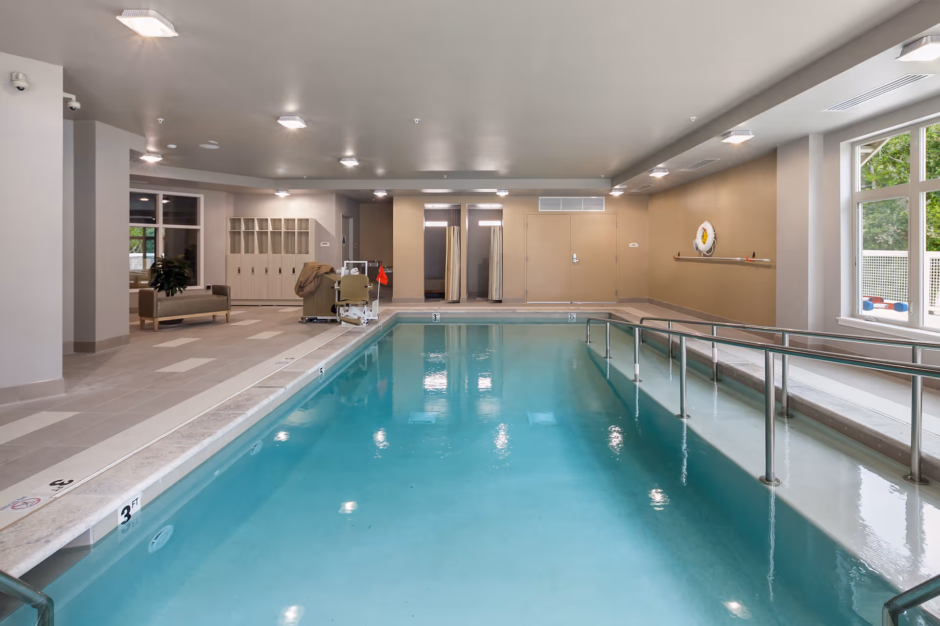 Indoor swimming pool with clear blue water, metal handrails, and a pool lift chair. The pool area has beige tiled flooring, lockers, a bench, and large windows letting in natural light. The walls are painted in neutral tones and there are ceiling lights illuminating the space.