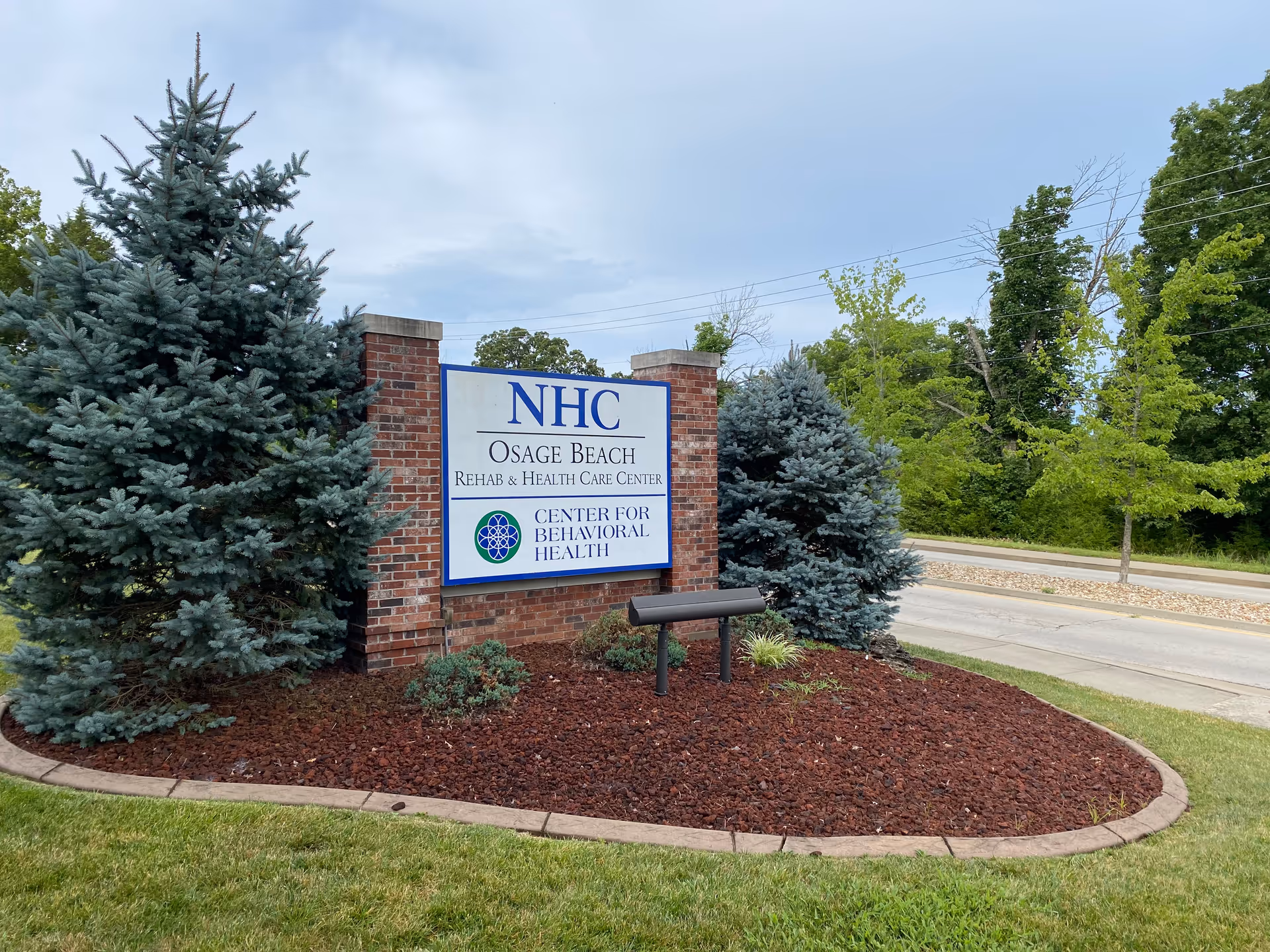 Brick entrance sign reading 'NHC Osage Beach Rehab & Health Care Center' with landscaping, evergreen trees, and a road in the background.
