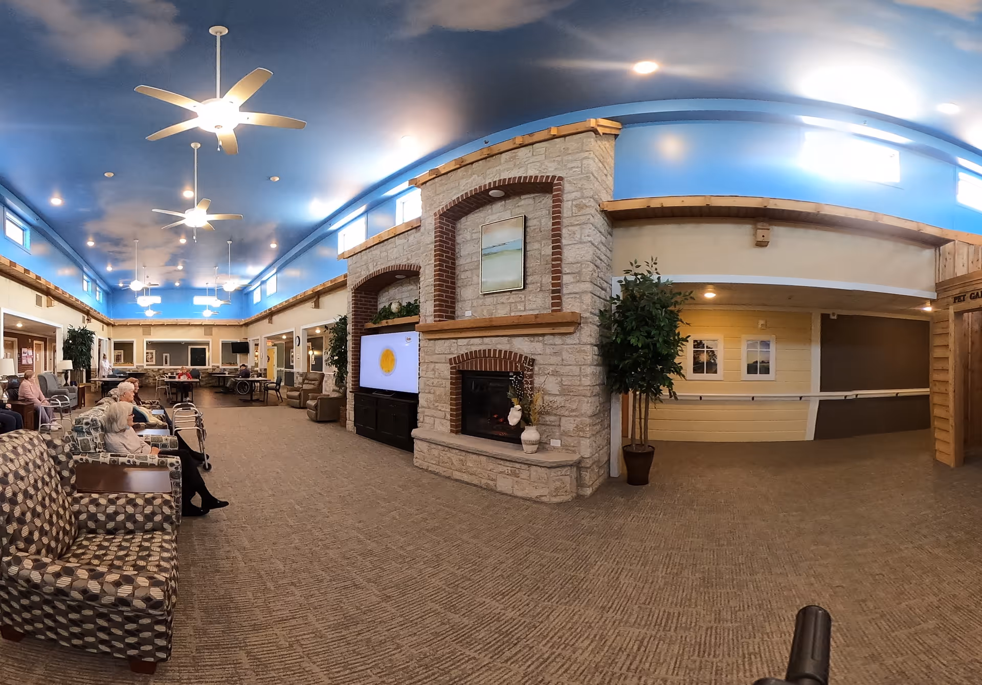 Spacious senior living community common lounge with seating, a stone fireplace and TV under a high blue ceiling.