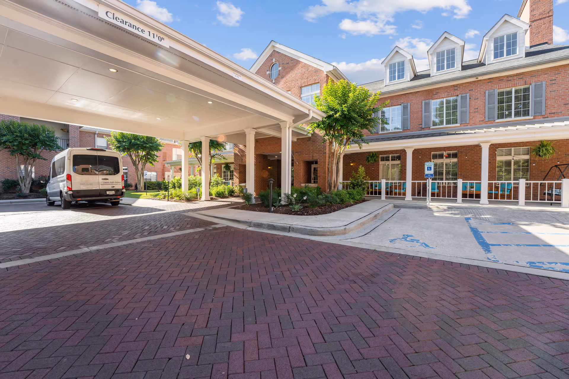 Entrance area of Town Village Audubon Park senior living facility showing a covered drop-off zone with clearance sign, a white van parked on the left, brick-paved driveway, accessible parking spaces, and a multi-story brick building with windows and greenery.