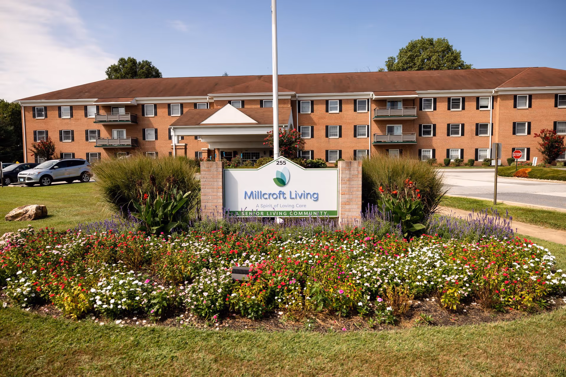 Front exterior view of Millcroft Living, a senior living community, showing a three-story brick building with balconies, a covered entrance, a well-maintained flower bed with colorful flowers, and a sign with the facility name in front.