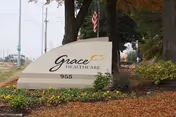 Outdoor view of a sign for Grace Healthcare at 955, surrounded by landscaping with flowers, mulch, and trees, with a street and utility poles visible in the background.