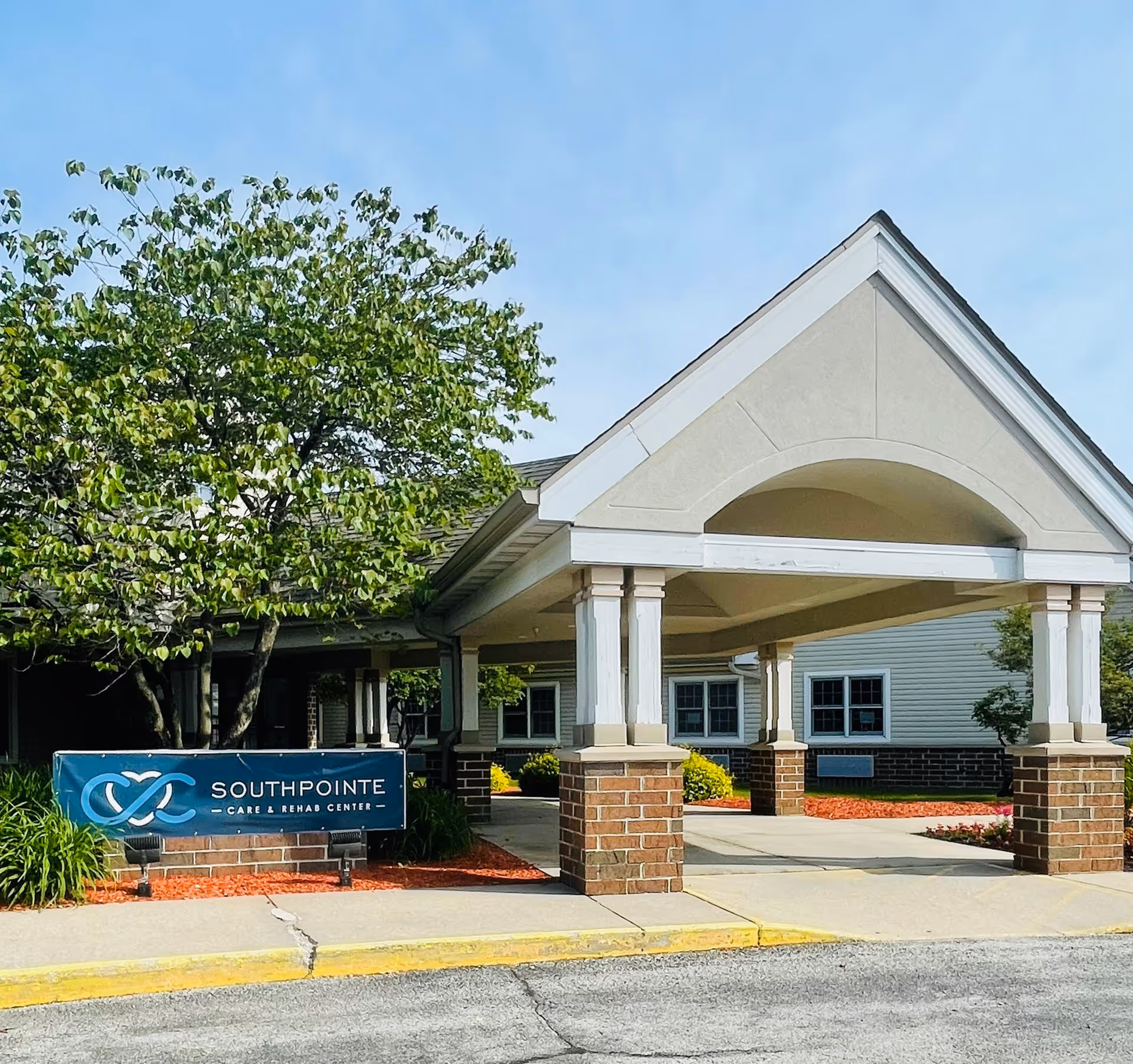 Exterior view of Southpointe Care & Rehab Center showing the entrance with a covered driveway supported by brick and white columns, a tree with green leaves, and a blue sign with the facility's name.