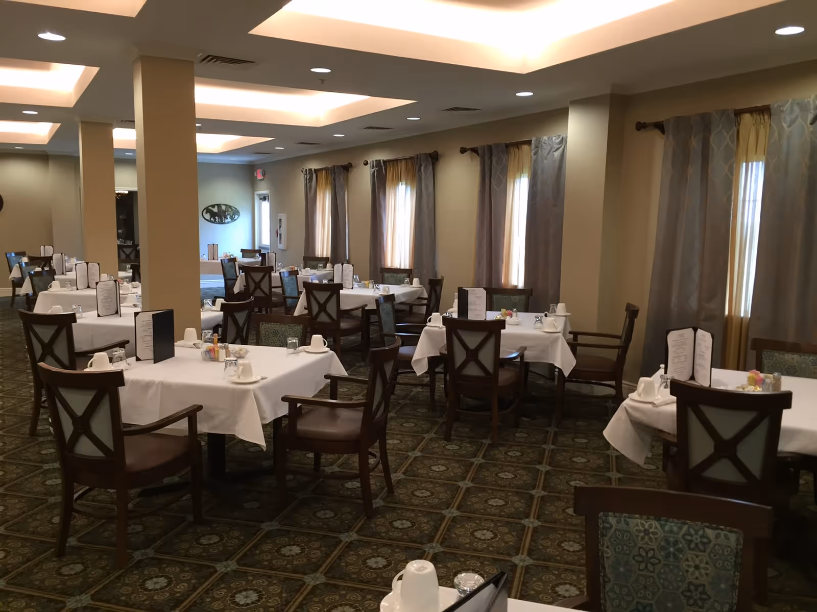 Interior view of a dining room with multiple tables covered with white tablecloths, each set with menus, cups, glasses, and condiments. The room has patterned carpet flooring, beige walls, and windows with curtains. Ceiling lights provide soft illumination.