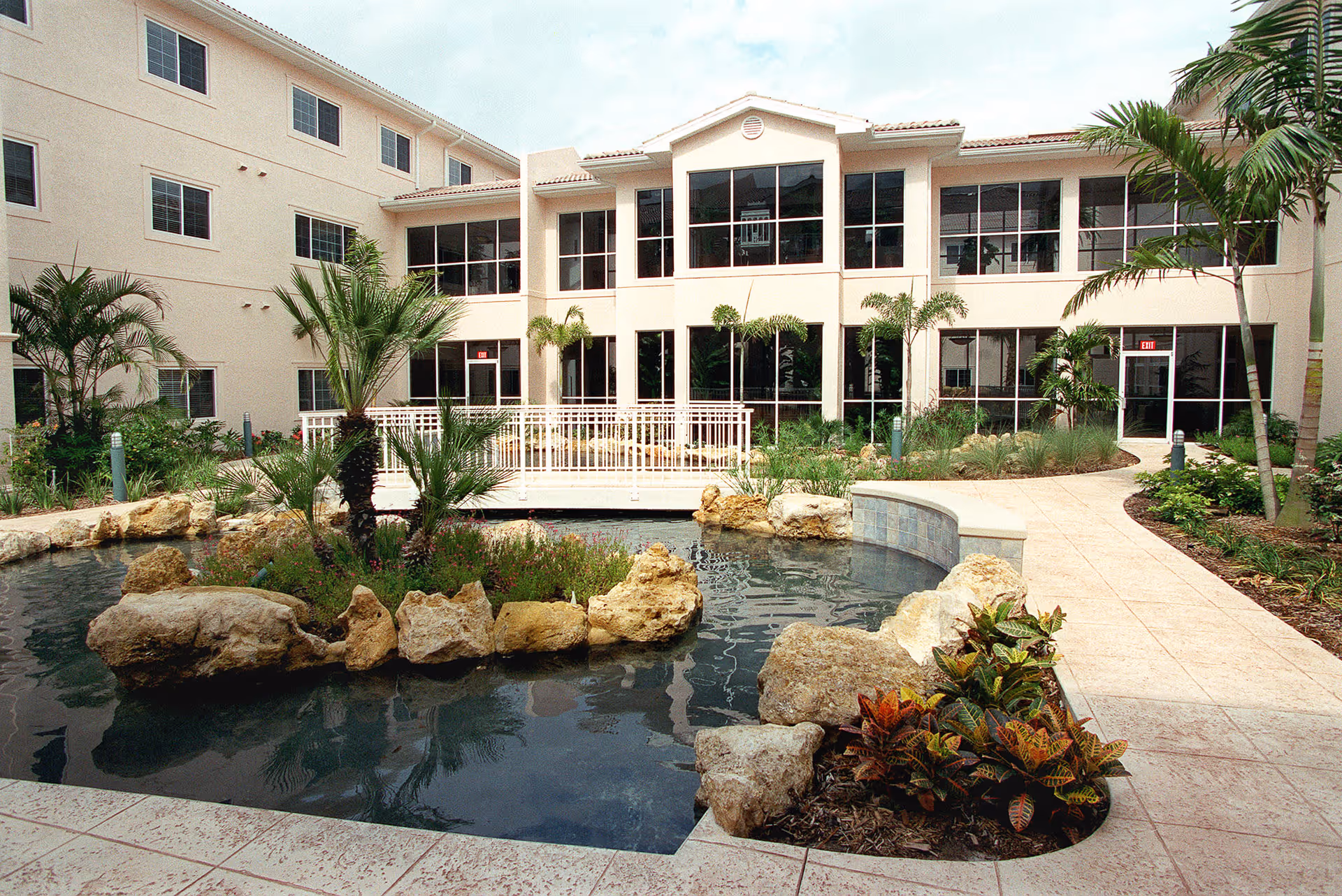 Sunlit courtyard with a decorative pond, rock landscaping, palm trees, a tiled walkway, and a two-story building with large windows.