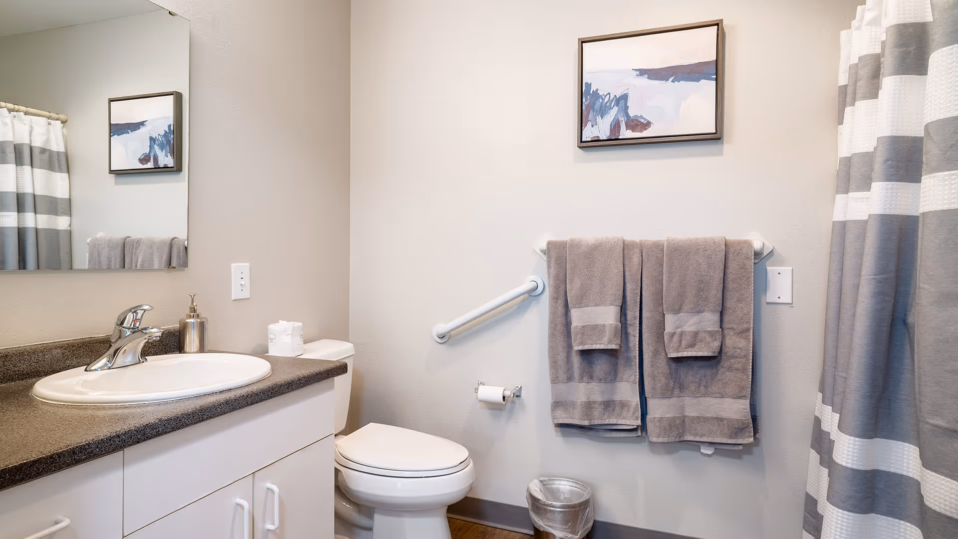 A clean and modern bathroom with a white sink and dark countertop, a toilet, a towel rack with two gray towels, a small trash can, a striped gray and white shower curtain, and a framed abstract painting on the wall.